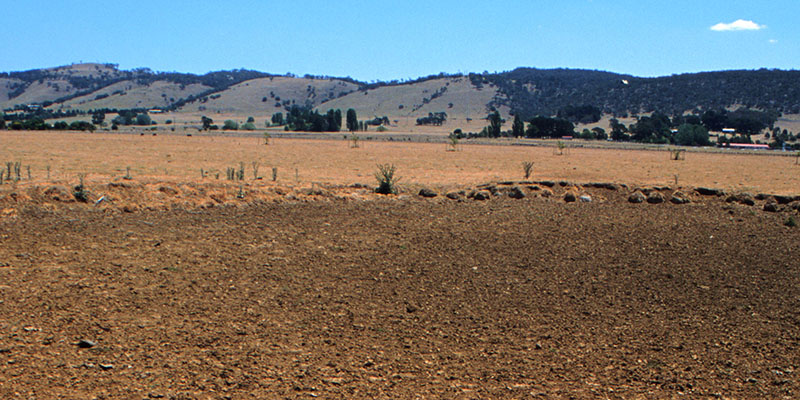 A view of a barren, dry landscape with a foreground of cracked earth and the background showing gently rolling hills under a clear blue sky.