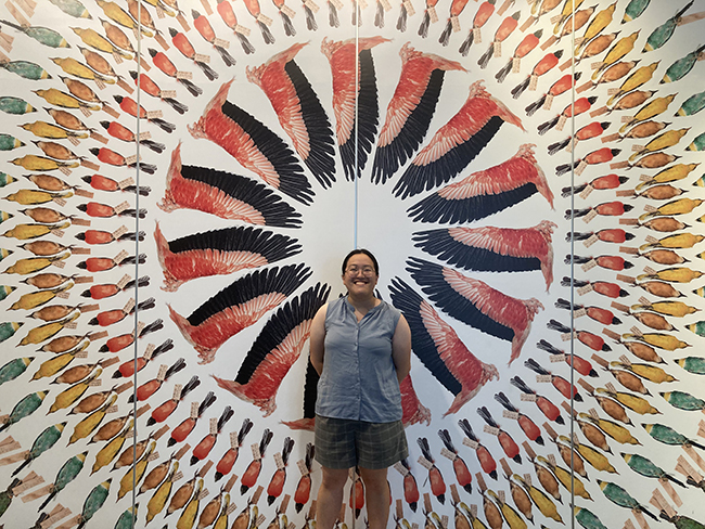 A woman smiling in front of a colorful wall display featuring a radiating pattern of feather illustrations.
