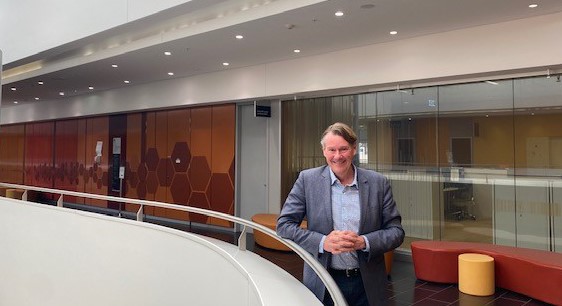 A man in business attire smiles while standing in a modern office space with orange and white decor.
