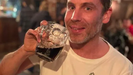 A man smiling while holding a large glass of dark beer in a lively indoor setting.