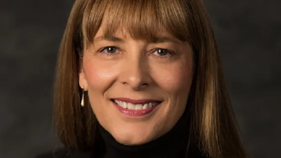 A portrait of a smiling woman with shoulder-length brown hair, wearing a black turtleneck and a gold necklace.