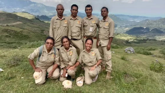 Group of seven park rangers in uniform posing together on a grassy hill with a scenic mountainous background.
