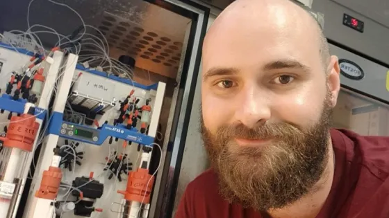 A man with a beard smiling in a laboratory next to equipment with various tubes and devices.