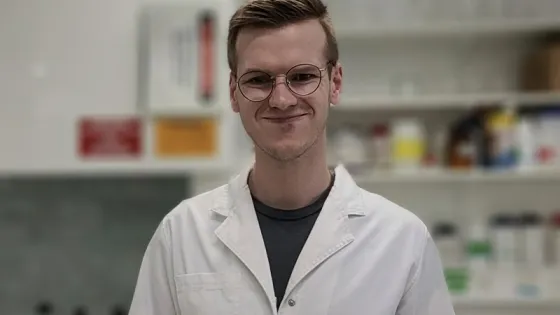 A young man with glasses wearing a white lab coat smiling in a laboratory setting.