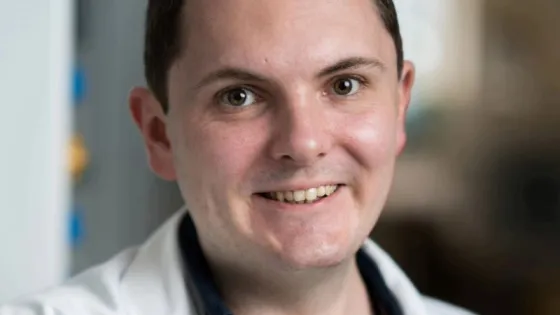 A smiling man wearing a white chef's coat stands in an indoor setting.