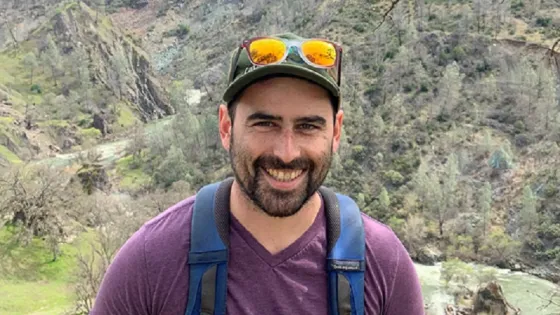 A man smiling at the camera while wearing a backpack and sunglasses, standing in front of a scenic mountainous landscape.