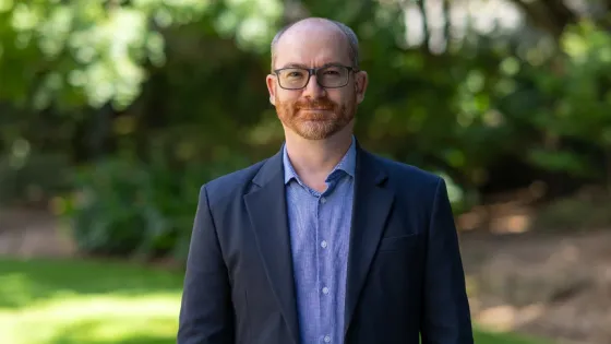 A bald man with glasses, wearing a navy blue suit jacket and a checked shirt, smiling at the camera with a green, leafy background.