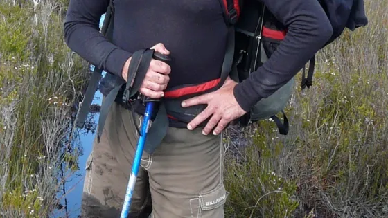 A male hiker with a large backpack and walking stick stands in a wetland area, wearing a hat and mud-covered boots.