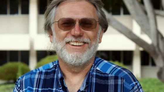 An older man with a gray beard and glasses, smiling in a blue plaid shirt, standing in front of a building with trees.