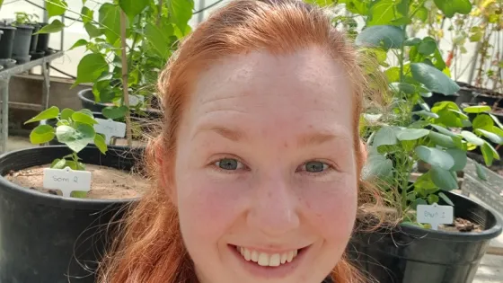 A woman with red hair smiling in a greenhouse surrounded by plants.