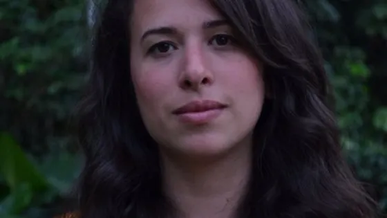 A woman with long dark hair wearing a mustard-colored top, standing in front of a green, leafy background.