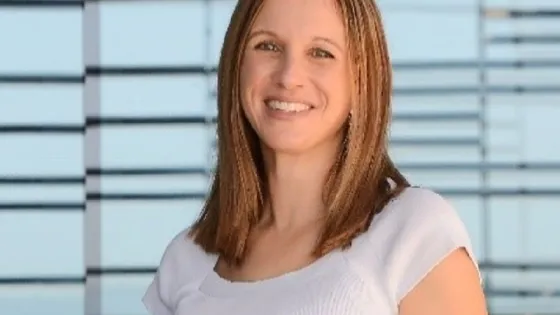 A smiling woman with shoulder-length brown hair, wearing a white top, stands in front of a blue glass building.