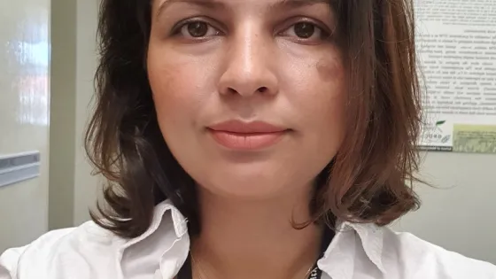 A portrait of a woman with shoulder-length hair wearing a white shirt, multiple necklaces, and a name badge, standing in an office.
