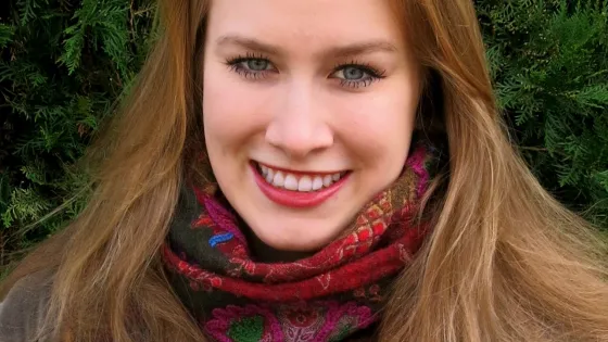 A woman with long blonde hair, smiling, wearing a colorful scarf, in front of a green bush.