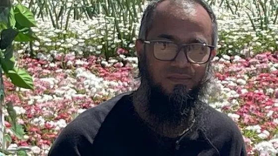 A man standing in front of a field of blooming flowers, wearing glasses and a black shirt.
