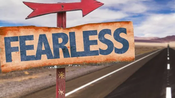 A weathered sign with the word "FEARLESS" pointing left beside a long, straight road through a desert landscape.
