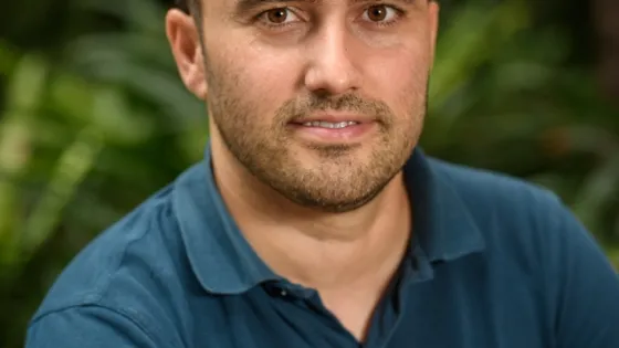 Portrait of a man in a teal polo shirt, smiling gently with a blurred green foliage background.