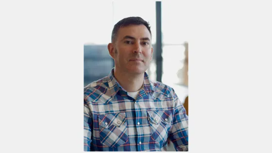 A man in a plaid shirt sitting indoors with a blurred background.