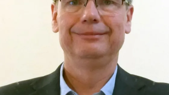 A man with glasses, wearing a suit jacket and a light blue shirt, smiles slightly at the camera.