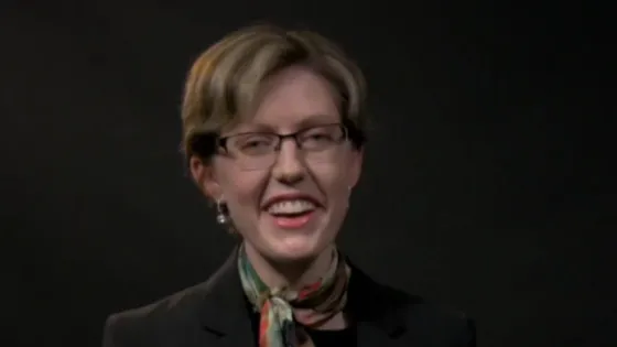 A woman with short hair and glasses, smiling broadly, wearing a blazer and a colorful scarf, against a dark background.
