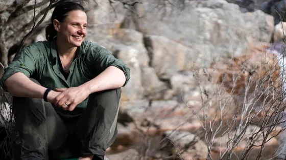 A person sits smiling on a rock in a natural, rocky landscape, surrounded by bare tree branches.
