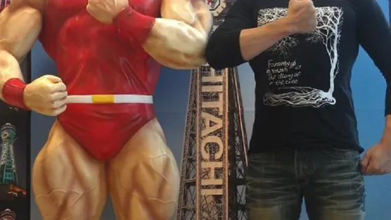 A person stands next to a large, muscular, red-suited statue, playfully mimicking its pose.