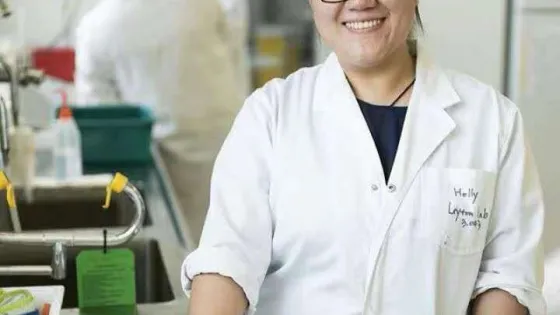 A smiling woman in a lab coat stands in a laboratory setting.