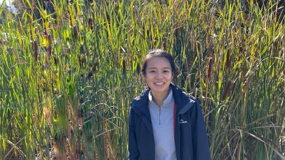 A young woman smiles in front of tall green and brown reeds under bright sunlight.