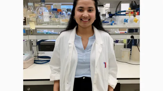 A woman in a white lab coat smiling in a laboratory with shelves stocked with colorful lab supplies in the background.