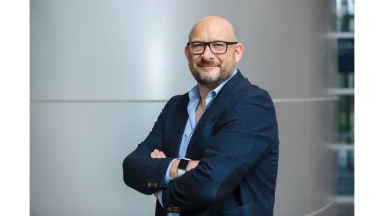 A bald man with glasses, wearing a blue suit and light blue shirt, stands smiling with his arms crossed in a modern office environment.
