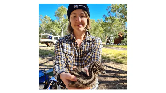 A woman in a plaid shirt and beanie holds a small emu chick outdoors.