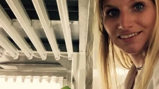 A woman in a lab coat smiling at the camera next to a small potted plant, with fluorescent lights above.
