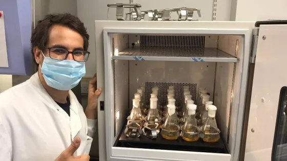 A scientist in a lab coat and mask gives a thumbs-up next to an open incubator containing several flasks of liquid.