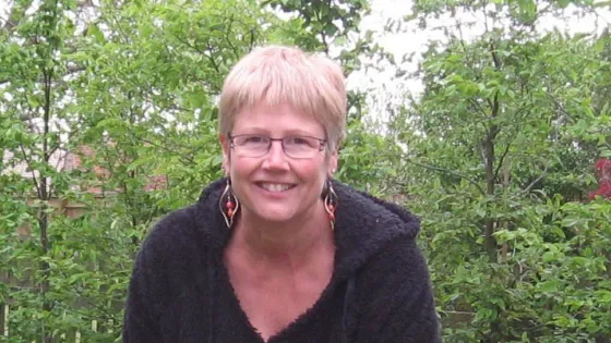 A smiling woman with short blonde hair and glasses, wearing a black sweater, seated outdoors with a background of lush greenery.