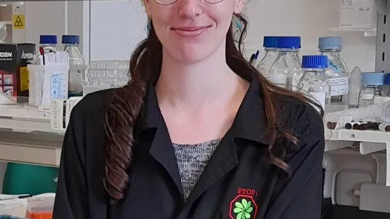 A woman with glasses and a ponytail, wearing a lab coat, smiles in a laboratory setting.