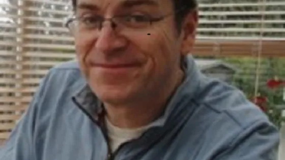 A man with glasses smiling and wearing a blue shirt, seated indoors with a white window blind background.