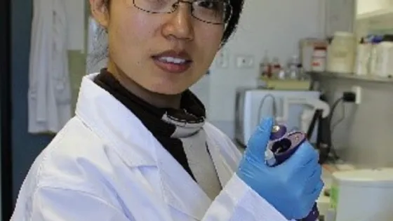 A scientist wearing a lab coat and gloves holds a test tube while standing in a laboratory.