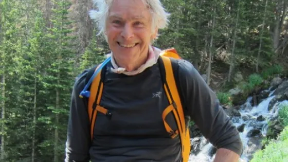 An elderly man with a joyful expression wearing a backpack stands in a forest near a stream.