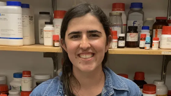 A woman smiling in a laboratory with shelves of chemical bottles behind her.