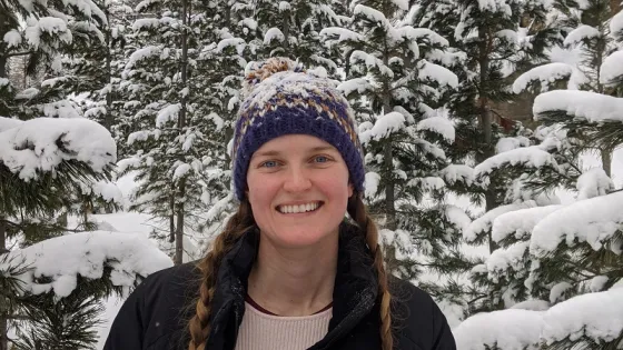 A woman in a winter hat and jacket smiles in front of snow-covered pine trees.