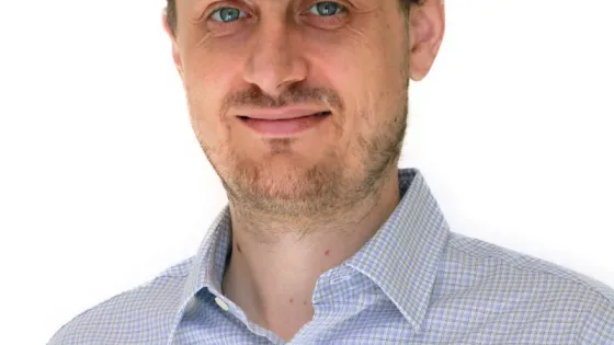 A man with short brown hair and a light beard, wearing a blue and white checkered shirt, smiling at the camera.
