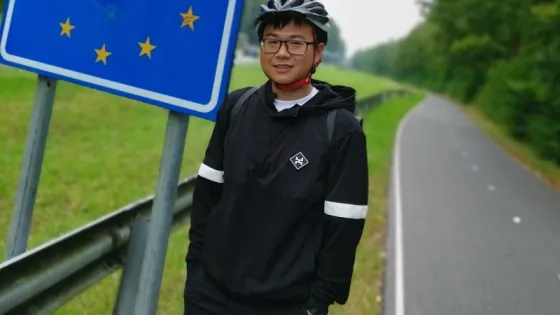 A person in cycling gear smiling next to a "Nederland" sign with a blue background and yellow stars.
