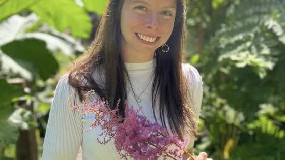 A woman smiling, holding pink flowers while standing in a lush garden.