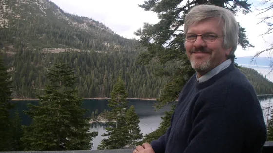 Man with glasses and a beard smiles while leaning on a railing, with a scenic view of a lake and pine-covered mountains in the background.