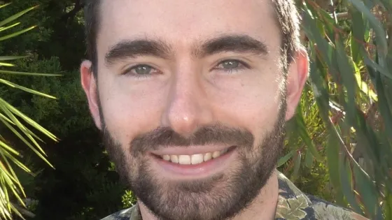 A man with a beard smiling in front of greenery, wearing a floral shirt.