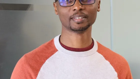 A man wearing glasses and a color-blocked sweater standing indoors and smiling at the camera.