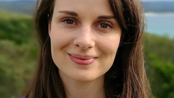 A portrait of a woman with long dark hair and blue eyes, smiling gently in front of a blurred natural landscape.
