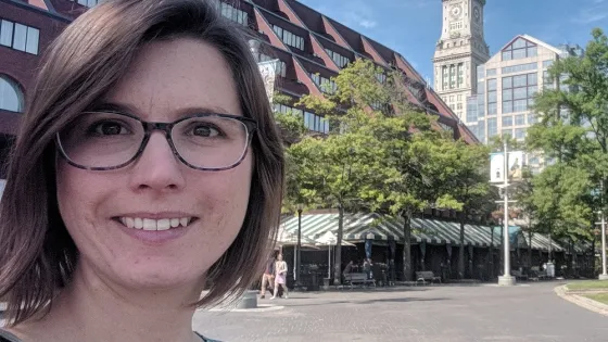 A woman with glasses smiling at the camera, with a historic clock tower and a modern building in the background on a sunny day.