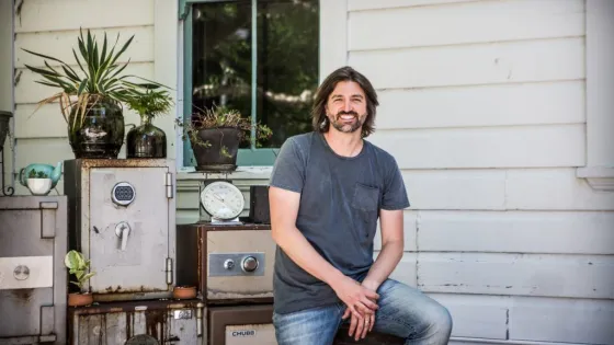 A man with long hair and a beard smiling while sitting on a stool in front of a vintage house, surrounded by old safes and scales with potted plants on top.
