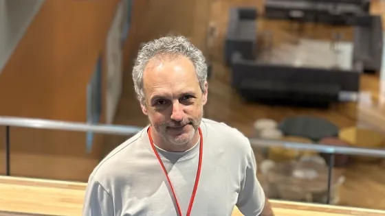 A man with graying hair, wearing a light gray T-shirt and black pants, stands leaning on a railing with an office space visible in the background.